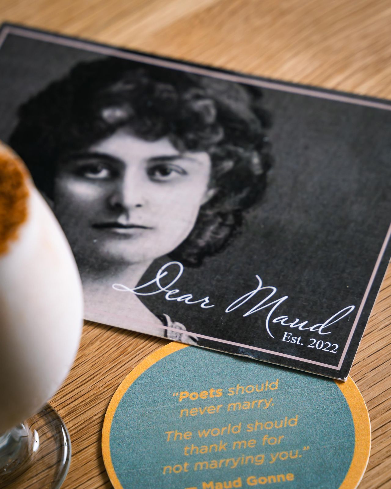 A Dear Maud table card with a portrait of Maud Gonne and the Est. 2022 mark, beside a cocktail
